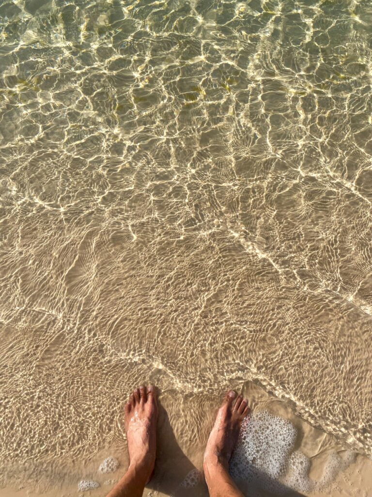 pexels-photo-27990192-27990192 Sunlit feet submerged in crystal clear ocean water at a tranquil sandy beach.