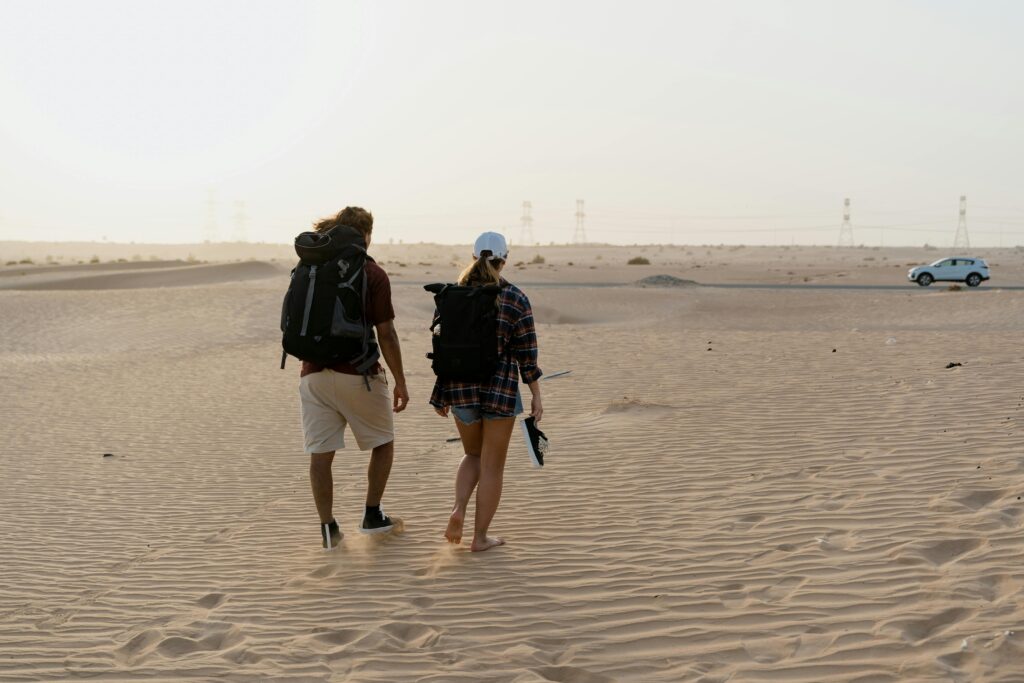 Two hikers walking through a vast desert landscape with a distant car and power lines.