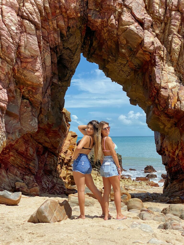 pexels-photo-12936082-12936082 Two women in denim shorts pose by a rock arch on a sunny Brazilian beach.
