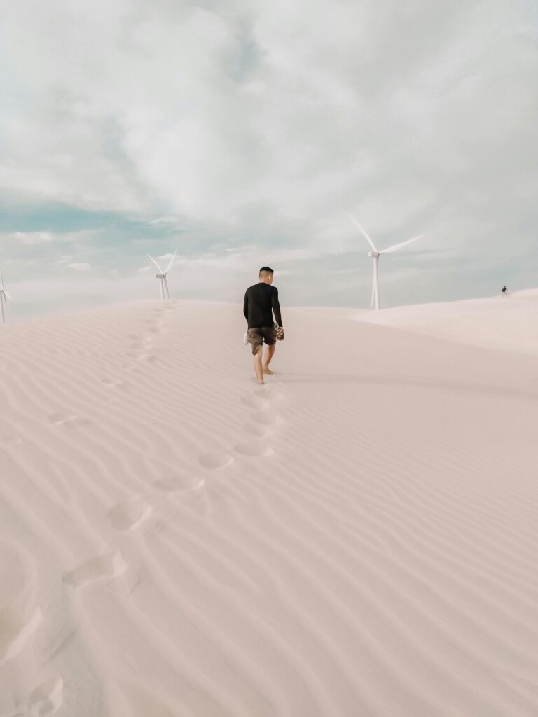 A person walks across Santo Amaro do Maranhão's sandy dunes with wind turbines in a tranquil landscape.