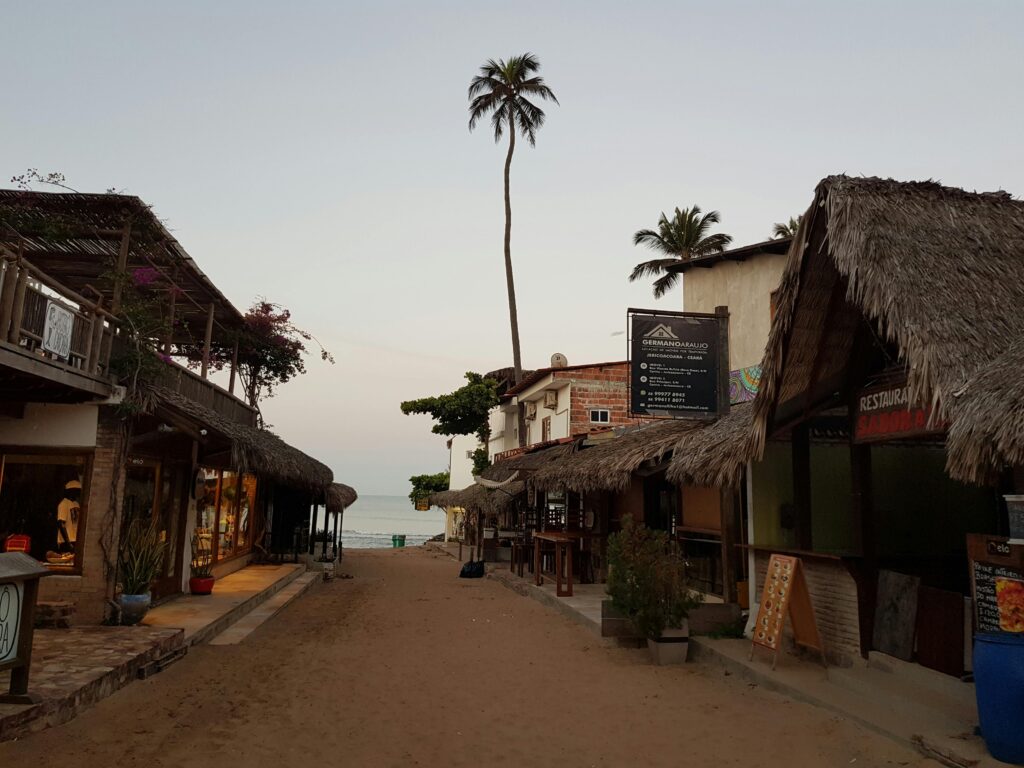 Tranquil street scene in Jericoacoara, Brazil, featuring palm-lined shops and thatched roofs at dusk.