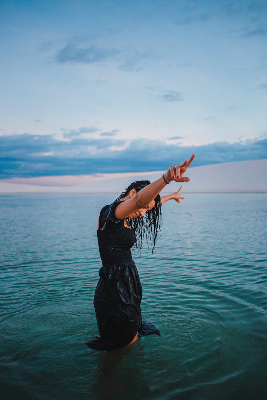 Woman in a black dress standing in water, posing with arms raised under a vibrant sunset sky.