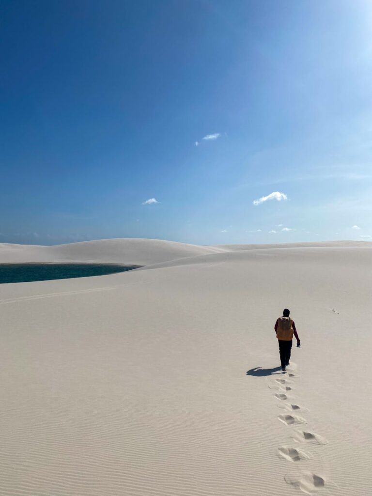 A lone traveler walks through the sandy dunes of Lençóis Maranhenses with a serene blue sky overhead.