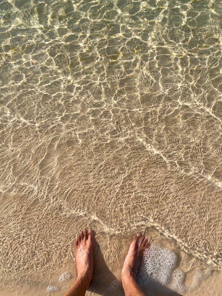 Sunlit feet submerged in crystal clear ocean water at a tranquil sandy beach.